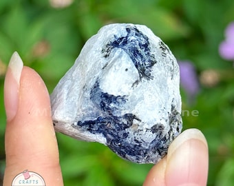 May include: A raw, natural stone with a white base and dark blue veining. The stone is held between fingers against a blurred green and purple background. The stone has a rough, textured surface.