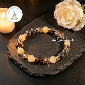 May include: A beaded bracelet featuring a mix of amber, clear, and dark gray stones, interspersed with gold-colored metal beads. The bracelet is displayed on a dark, textured surface, with the text "find your spark". A lit candle and a white rose are in the background.