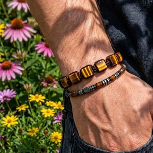 May include: Two bracelets worn together. The top bracelet features square, golden-brown tiger's eye beads on a dark cord. The bottom bracelet consists of alternating small, metallic silver-toned beads and brown tiger's eye beads on a thin cord.
