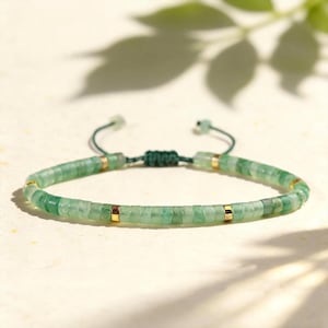 May include: A bracelet featuring small, flat, circular green stone beads arranged in a continuous row, accented by four evenly spaced gold-toned metal spacers. The piece includes a dark green adjustable cord closure with a sliding knot mechanism.