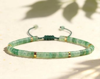 May include: A bracelet featuring small, flat, circular green stone beads arranged in a continuous row, accented by four evenly spaced gold-toned metal spacers. The piece includes a dark green adjustable cord closure with a sliding knot mechanism.