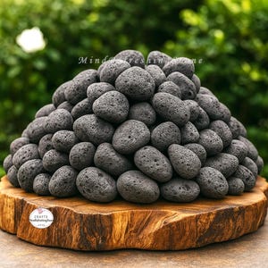 Op de afbeelding: Een stapel donkergrijze, poreuze lavastenen gerangschikt op een natuurlijke houten plaat. De stenen zijn van verschillende grootte en vorm, waardoor een textuurrijke, organische weergave ontstaat. De houten plaat heeft een ruwe, natuurlijke rand. De tekst "Mind Freshing Stone" is zichtbaar.