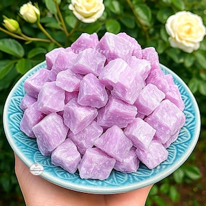 Puede incluir: Un plato de cerámica turquesa rebosante de cristales morados claros. Los cristales tienen forma de cubo y un aspecto ligeramente translúcido. El plato se sostiene frente a un fondo de follaje verde y rosas amarillas.