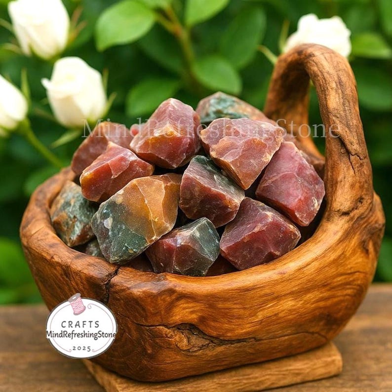 May include: A wooden basket filled with raw, colorful gemstones. The stones are a mix of red, green, and brown hues. The basket has a natural wood grain and a handle. The background features white flowers and green foliage. The text "MindRefreshingStone" is visible.