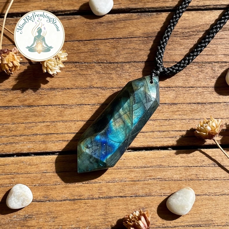 May include: A faceted, elongated labradorite pendant necklace with a black cord. The gemstone displays iridescent blue and green hues. The necklace is set against a wooden surface, with small white stones and dried flowers scattered around.