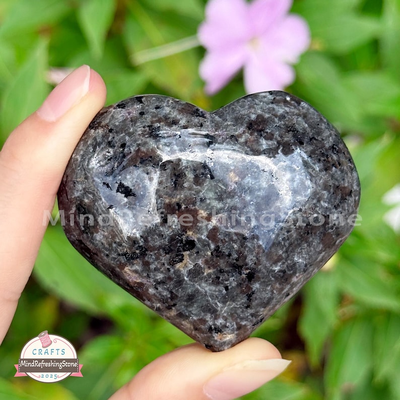 Puede incluir: Una piedra en forma de coraz&oacute;n con un patr&oacute;n moteado gris oscuro y negro. La piedra se sostiene en una mano, con un fondo borroso de follaje verde y una flor rosa. El texto "Mind Refreshing Stone" es visible.