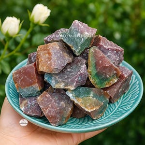 May include: A collection of raw, colorful gemstones in shades of red, green, purple, and brown, displayed in a teal-colored bowl. The rough-cut stones have a natural, textured appearance, with variations in color and shape. The bowl is held in a hand.