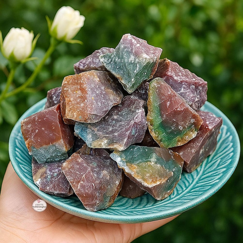 May include: A collection of raw, colorful gemstones in shades of red, green, purple, and brown, displayed in a teal-colored bowl. The rough-cut stones have a natural, textured appearance, with variations in color and shape. The bowl is held in a hand.