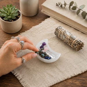 May include: A white crescent moon-shaped dish containing a collection of small polished gemstones including purple, pink, clear, and dark grey stones, positioned near a bundle of dried sage tied with twine.