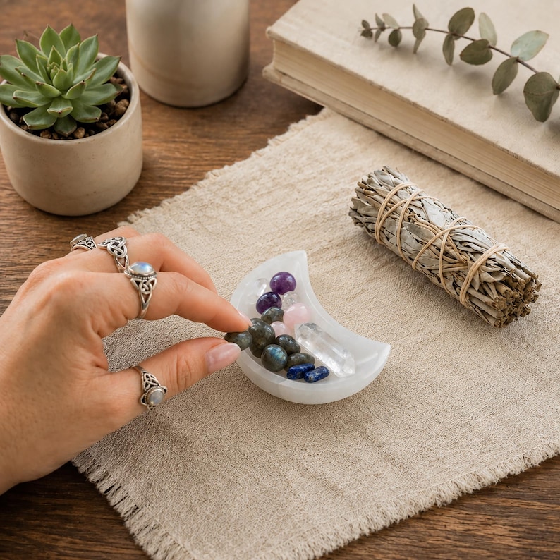 May include: A white crescent moon-shaped dish containing a collection of small polished gemstones including purple, pink, clear, and dark grey stones, positioned near a bundle of dried sage tied with twine.