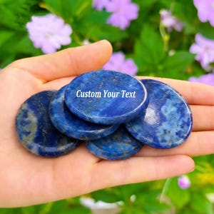 May include: A collection of five smooth, polished, circular lapis lazuli stones with deep blue coloring and natural white and gold flecks. The top stone features the white text Custom Your Text printed clearly across its center surface.