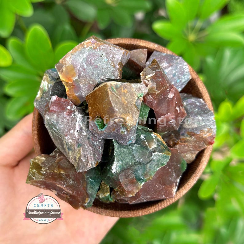 May include: A wooden bowl filled with raw, uncut gemstones. The stones are a mix of colors, including green, brown, and red. The bowl is held in a hand, with green foliage in the background. The image includes a logo that says "CRAFTS MindRefreshingStone 2025".