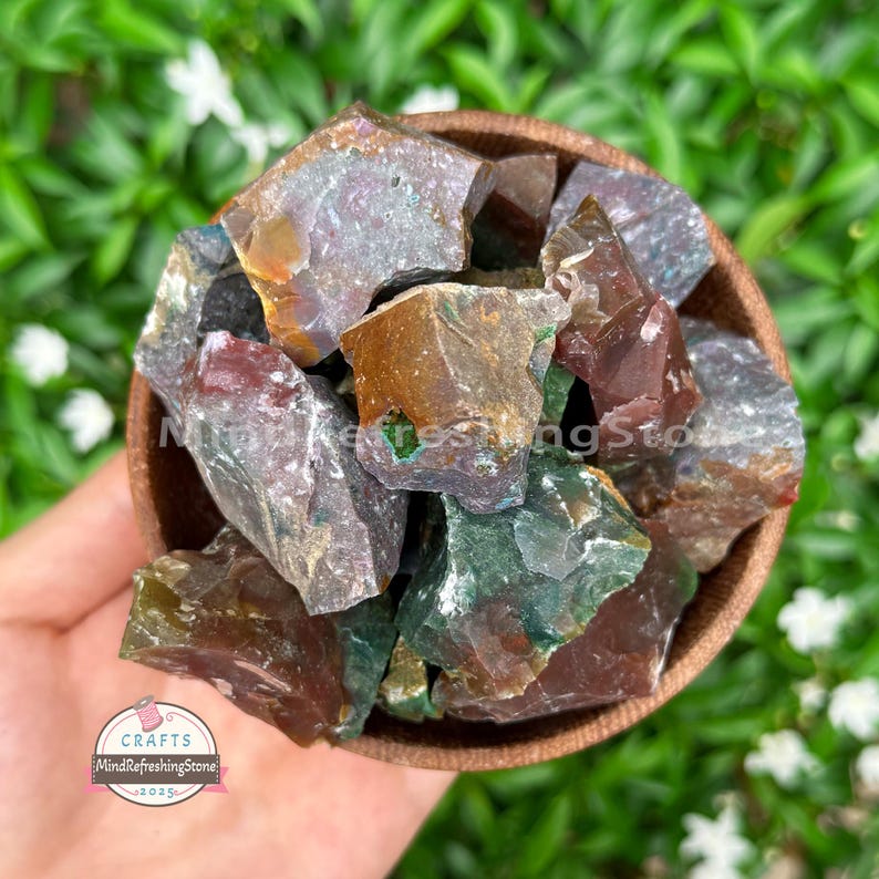 May include: A wooden bowl filled with raw, uncut gemstones. The stones display a variety of colors, including shades of green, brown, and red. The bowl is held in a hand, with a blurred green background.
