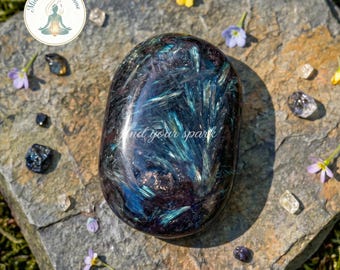 Puede incluir: Una piedra pulida de forma ovalada con tonos negro profundo y morado oscuro, con inclusiones vibrantes e iridiscentes de color azul en forma de aguja. La superficie muestra el texto "find your spark" en una delicada fuente cursiva blanca.