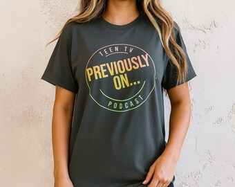 Op de afbeelding: Donkergrijs T-shirt met een cirkelvormige afbeelding met de tekst "TEEN TV PREVIOUSLY ON... PODCAST" in een roze, gele en groene gradiënt. Het shirt heeft korte mouwen en een ronde hals.