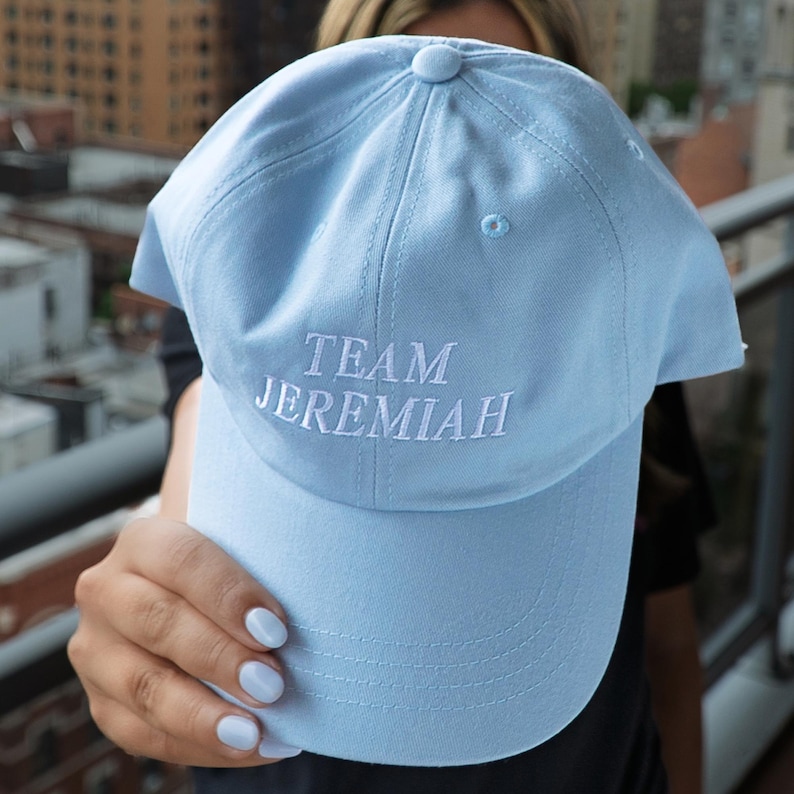 May include: A light blue baseball cap with the embroidered text "TEAM JEREMIAH" in white. The cap has a curved brim and a button on top. The cap is made of a soft fabric.