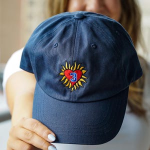 May include: Navy blue baseball cap with a curved brim. The cap features an embroidered design of a red heart with a yellow sunburst and the number 3 in blue. The cap is made of a soft, durable fabric.
