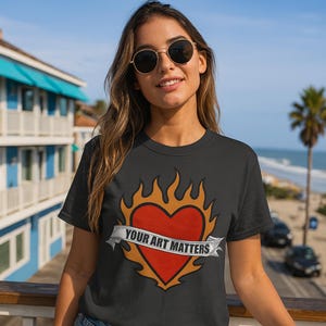 May include: Black t-shirt featuring a red heart design with orange flames and a banner that reads "YOUR ART MATTERS." The shirt is a crew neck style and is being worn outdoors.