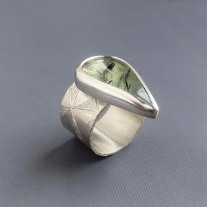 May include: A silver ring with a wide band featuring a geometric pattern. The ring has a teardrop-shaped gemstone with green and black inclusions. The gemstone is set in a bezel setting, and the ring is displayed against a gray background.