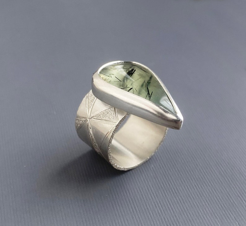 May include: A silver ring with a wide band featuring a geometric pattern. The ring has a teardrop-shaped gemstone with green and black inclusions. The gemstone is set in a bezel setting, and the ring is displayed against a gray background.