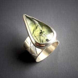May include: A silver ring featuring a large, pear-shaped green gemstone with dark inclusions. The ring's band has a textured, geometric design. The gemstone is set in a polished silver bezel, creating a striking contrast.