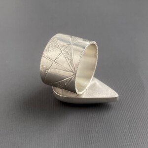 May include: A silver ring with a geometric design. The ring features a wide band with a textured pattern of triangles and lines. The ring's base has a smooth, teardrop shape. The ring is displayed against a gray background.