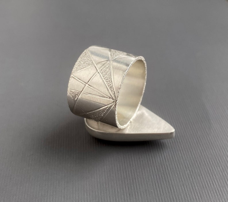May include: A silver ring with a geometric design. The ring features a wide band with a textured pattern of triangles and lines. The ring's base has a smooth, teardrop shape. The ring is displayed against a gray background.