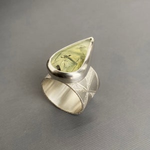May include: A silver ring featuring a large, teardrop-shaped gemstone with a light green hue and black inclusions. The ring's band has a geometric starburst pattern and a textured edge, creating a unique and elegant design.
