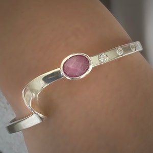 May include: A silver bangle bracelet featuring a pink oval gemstone and three smaller round clear gemstones.