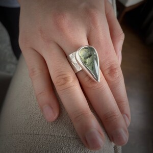 May include: A silver ring with a large, teardrop-shaped green gemstone. The ring has a wide band with etched starburst designs. The gemstone is a light green color with dark green inclusions, set in a polished silver bezel.