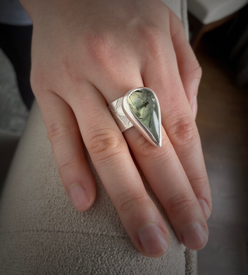 May include: A silver ring with a large, teardrop-shaped green gemstone. The ring has a wide band with etched starburst designs. The gemstone is a light green color with dark green inclusions, set in a polished silver bezel.