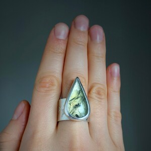 May include: A silver ring featuring a large, teardrop-shaped gemstone with green and black inclusions. The ring has a wide band with a textured design. The gemstone is set in a silver bezel, creating a striking contrast.