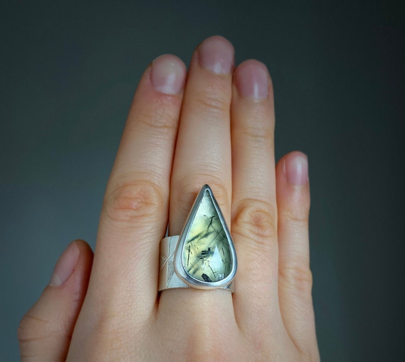 May include: A silver ring featuring a large, teardrop-shaped gemstone with green and black inclusions. The ring has a wide band with a textured design. The gemstone is set in a silver bezel, creating a striking contrast.