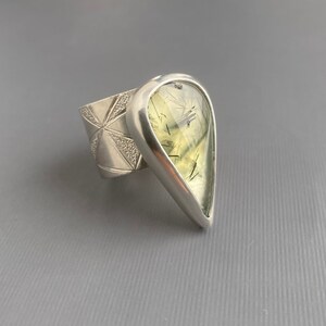 May include: A silver ring featuring a large, pear-shaped gemstone with a pale green hue and dark inclusions. The ring's band has a geometric design. The gemstone is set in a polished silver bezel.