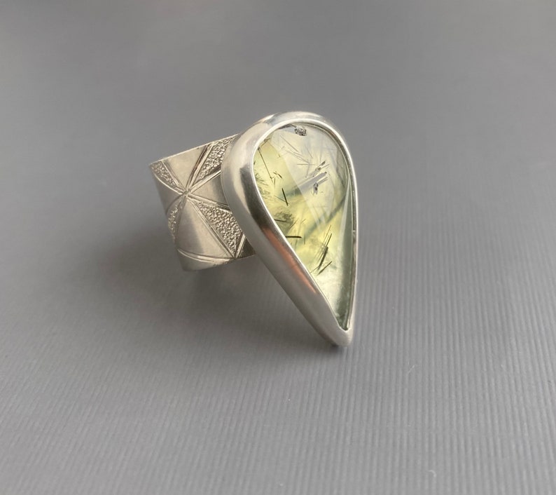 May include: A silver ring featuring a large, pear-shaped gemstone with a pale green hue and dark inclusions. The ring's band has a geometric design. The gemstone is set in a polished silver bezel.