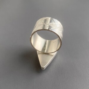 May include: A silver ring with a geometric design. The ring features a wide band with etched lines and a triangular base. The ring is made of a shiny, metallic material and is set against a gray background.