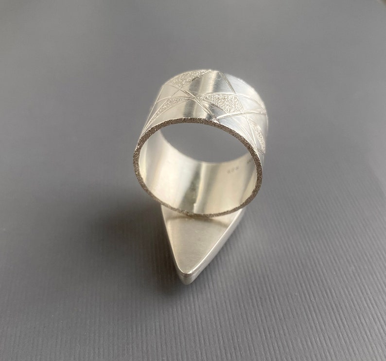 May include: A silver ring with a geometric design. The ring features a wide band with etched lines and a triangular base. The ring is made of a shiny, metallic material and is set against a gray background.
