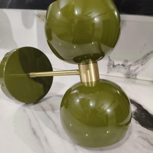 May include: Olive green modern wall sconce with two spherical shades and a circular base. The fixture has a brass arm and accents, creating a stylish contrast. The glossy finish reflects light, adding a contemporary touch to any interior space.