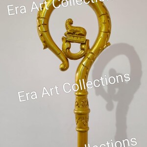 May include: A gold-colored staff with a curved top and a detailed design. The top features a circular element with a lamb figure and decorative carvings. The staff has a long, straight shaft with ornate detailing. The text "Era Art Collections" is visible.