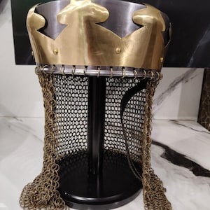 Könnte beinhalten: Ein mittelalterlicher Helm mit einer Messingkrone und Kettenrüstung. Die Krone hat ein dekoratives, spitzes Design. Der Helm wird auf einem schwarzen Ständer präsentiert. Die Kettenrüstung hängt vom Helm herunter.