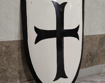 May include: A white shield with a black cross design. The shield has a black border with rivets. The cross is centered on the shield. The shield is a medieval or historical item.