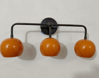 May include: A contemporary wall light with three orange globe shades. The fixture features a black metal frame and a circular base. The shades are evenly spaced, creating a warm glow. The light is designed for indoor use and would suit a modern home.