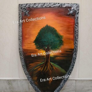 Op de afbeelding: Schildvormig kunstwerk met een boom met groene bladeren en wortels tegen een zonsondergang. Het kunstwerk heeft een getextureerde, zilverkleurige rand met decoratieve elementen. De tekst "Era Art Collections" is afgedrukt.