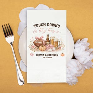 May include: A white rectangular paper napkin featuring the text TOUCH DOWNS & Tiny Toes, OLIVIA ANDERSON, and 08.20.2026. The graphic includes a football with a pink bow, a beer bottle, two beer mugs, baby shoes, and small pumpkins.