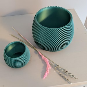 Könnte beinhalten: Zwei teal-farbene, gerippte, dekorative Pflanzgefäße in verschiedenen Größen. Der größere Pflanztopf ist oval geformt, der kleinere rund. Getrocknete rosa und graue dekorative Stiele sind zwischen den Pflanzgefäßen platziert. Die Pflanzgefäße stehen auf einer hellen Oberfläche.