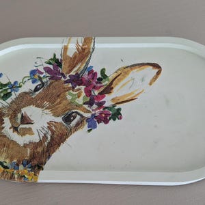 Könnte beinhalten: Ovale Dekoschale mit einem gemalten Bild eines braunen Hasen mit Blumenkranz. Die Schale hat einen hellbeigen Hintergrund und einen hellgrauen Rand. Das Fell des Hasen ist detailliert, und die Blumen sind in Lila, Blau und Gelb gehalten.