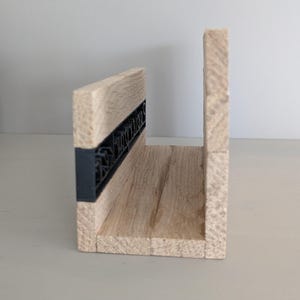 Könnte beinhalten: Ein U-förmiges Holzregal mit natürlicher Holzmaserung. Ein schwarzes, dekoratives Element ist vorne eingelassen. Das Regal ist für die Wandmontage konzipiert und eignet sich zur Präsentation kleiner Gegenstände.