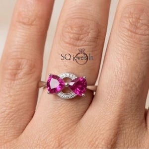 May include: A silver-toned ring featuring two pear-cut vibrant pink gemstones arranged in a bow shape, centered over a circular band encrusted with small clear stones. The text SQ jewels In is printed above the ring on the finger.