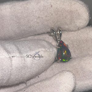 以下が含まれることがあります： 洋梨型の黒オパール宝石が付いたシルバーペンダント。オパールは赤、緑、黄色のきらめきを示しています。ペンダントは白い布の背景にあります。SQ jewels Inのジュエリーです。