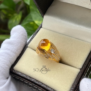Puede incluir: Un anillo de oro con una gran piedra preciosa ovalada de color ámbar en el centro. El anillo está incrustado con pequeñas piedras preciosas transparentes y presenta un diseño único. El anillo se presenta en una caja de joyería marrón con el texto "SQ jewels In".